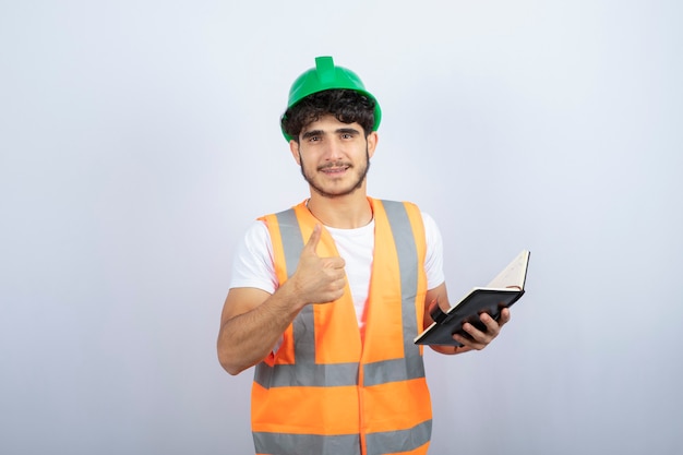 Junger männlicher Ingenieur im grünen Helm, der Notizbuch auf weißem Hintergrund hält. Hochwertiges Foto