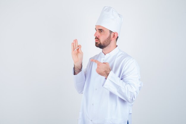 Junger männlicher Koch, der auf Zigarette in der weißen Uniform zeigt und nachdenklich aussieht. Vorderansicht.