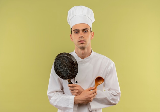 Kostenloses Foto junger männlicher koch, der kochuniform trägt, die bratpfanne und löffel in seiner hand auf isolierter grüner wand mit kopienraum kreuzt