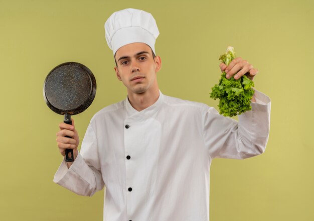 junger männlicher Koch, der Kochuniform trägt, die Bratpfanne und Salat in seiner Hand auf isolierter grüner Wand anhebt