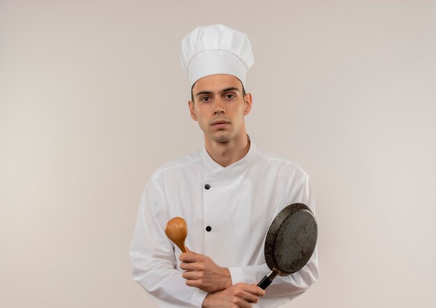 junger männlicher Koch, der Kochuniform trägt, die Hände hält, die Bratpfanne und Löffel auf isolierter weißer Wand mit Kopienraum halten