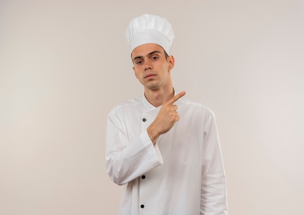 junger männlicher Koch, der Kochuniform trägt, zeigt Finger zur Seite auf isolierte weiße Wand mit Kopienraum