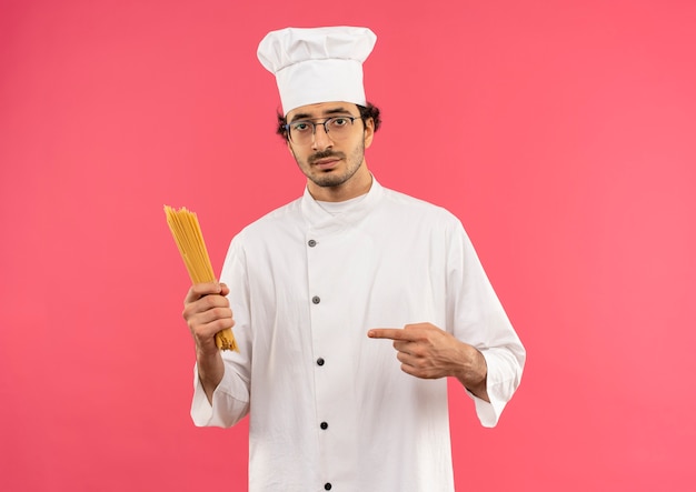 junger männlicher Koch, der Kochuniform und Brille hält und auf Spaghetti zeigt