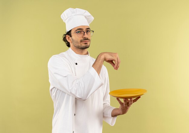 junger männlicher Koch, der Kochuniform und Gläser hält Platte und vorgibt, Salz zu verschütten