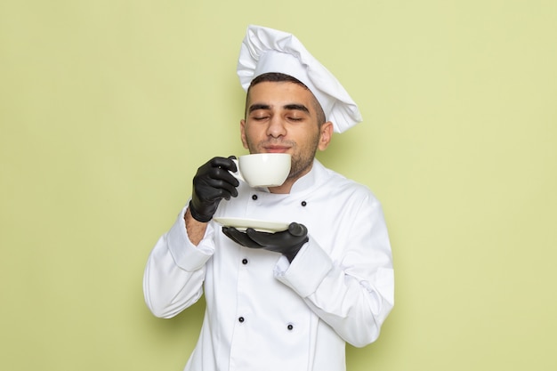 Junger männlicher Koch der Vorderansicht im weißen Kochanzug, der dunkle Handschuhe trägt und Kaffee auf Grün trinkt