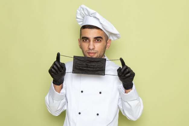 Junger männlicher Koch der Vorderansicht im weißen Kochanzug, der dunkle Handschuhe trägt und Maske auf Grün hält