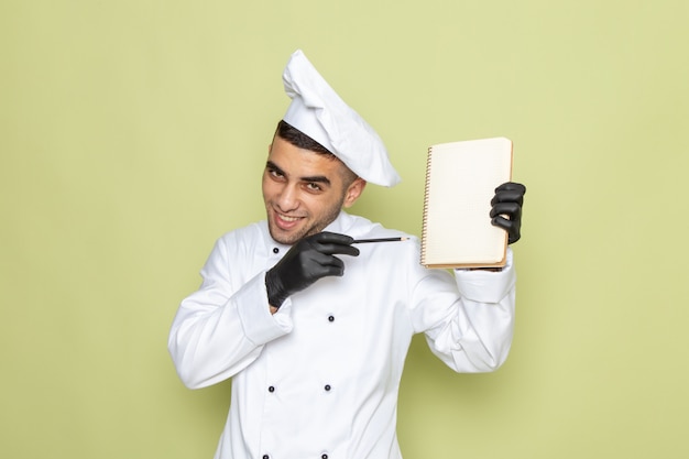 Junger männlicher Koch der Vorderansicht im weißen Kochanzug, der Notizblock hält und auf Grün lächelt