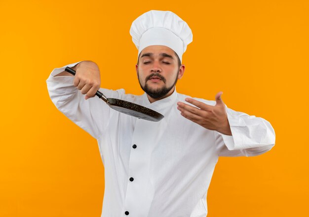 Junger männlicher Koch in der Kochuniform, die Bratpfanne hält und mit geschlossenen Augen schnüffelt und Hand auf Luft lokalisiert auf orange Raum