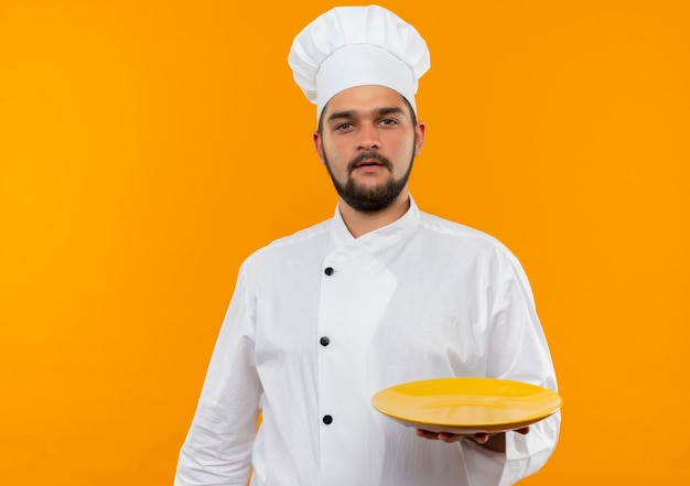 Junger männlicher Koch in der Kochuniform, die leere Platte hält, die auf orange Raum lokalisiert schaut