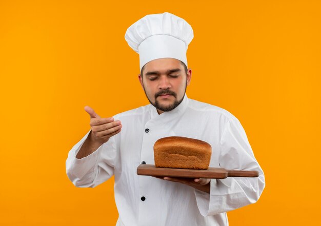 Junger männlicher Koch in der Kochuniform, die Schneidebrett mit Brot auf ihm hält und mit Hand auf Luft schnüffelt und Augen geschlossen auf orange Raum isoliert