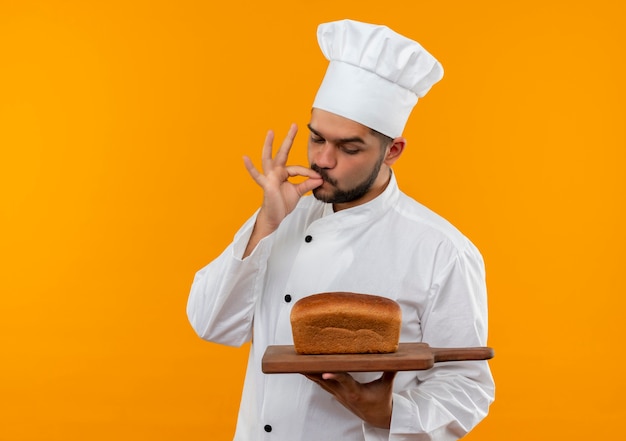 Junger männlicher Koch in der Kochuniform, die Schneidebrett mit Brot darauf hält und betrachtet und leckere Geste tut, die auf orange Raum lokalisiert wird