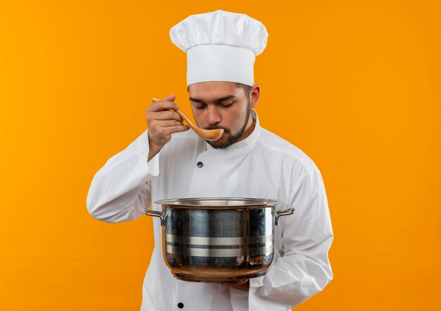 Junger männlicher Koch in der Kochuniform, die Topf hält und vom Löffel isst, der auf orange Raum lokalisiert wird