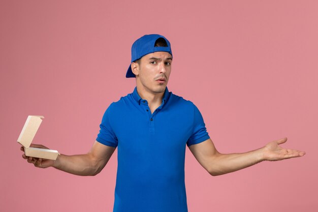 Junger männlicher Kurier der Vorderansicht im blauen Uniformumhang, der kleines leeres Liefernahrungsmittelpaket an der rosa Wand hält