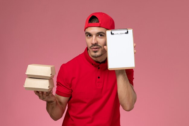 Junger männlicher Kurier der Vorderansicht im roten Uniformumhang, der kleine Pakete mit Essen und Notizblock auf hellrosa Hintergrund hält.