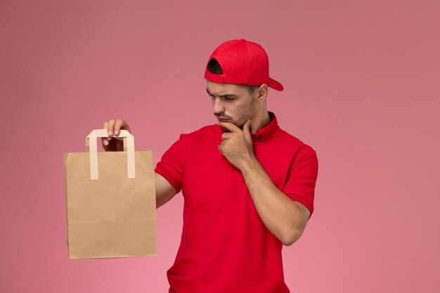 Junger männlicher Kurier der Vorderansicht im roten Uniformumhang, der Papiernahrungsmittelpaket hält, das auf dem hellrosa Hintergrund denkt.