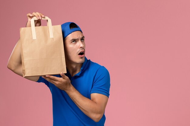 Junger männlicher Kurier der Vorderansicht in blauer Uniform und Umhang mit Lieferpapierpaket auf seinen Händen an der rosa Wand