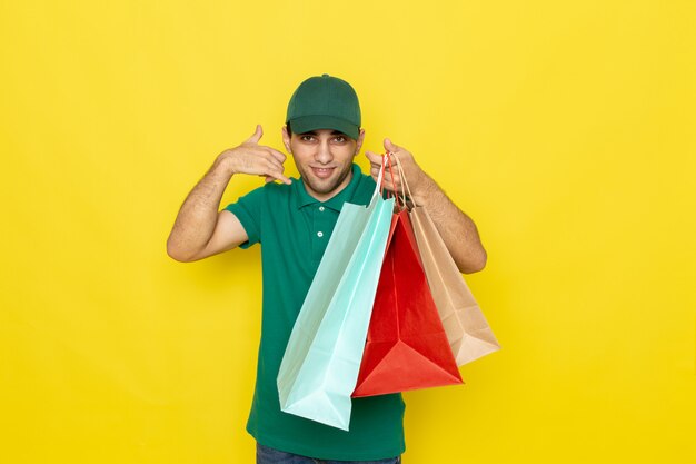Junger männlicher Kurier der Vorderansicht in der grünen Kappe des grünen Hemdes, die Einkaufspakete hält und Telefonanrufzeichen zeigt
