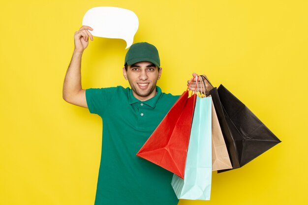 Junger männlicher Kurier der Vorderansicht in der grünen Kappe des grünen Hemdes, die Einkaufspakete und weißes Zeichen hält