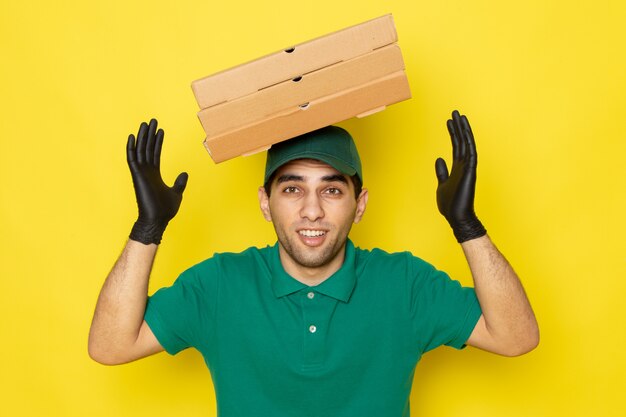 Junger männlicher Kurier der Vorderansicht in der grünen Kappe des grünen Hemdes, die Lieferkästen mit seinem Kopf in den Handschuhen auf Gelb hält