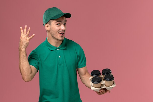 Junger männlicher Kurier der Vorderansicht in der grünen Uniform, die braune Kaffeetassen auf dem rosa Hintergrund hält