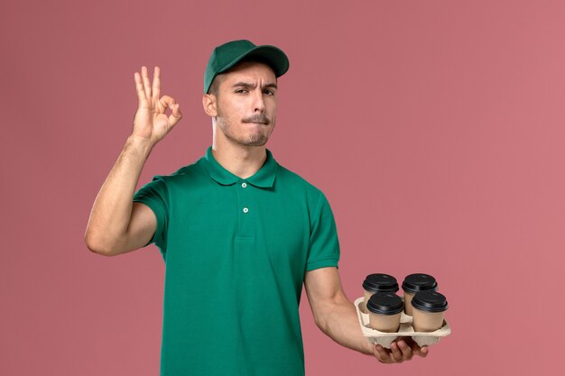 Junger männlicher Kurier der Vorderansicht in der grünen Uniform, die braune Kaffeetassen auf hellrosa Hintergrund hält