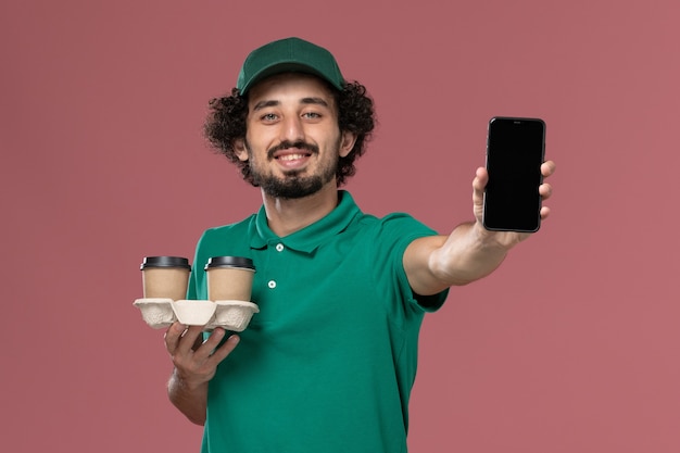 Junger männlicher Kurier der Vorderansicht in der grünen Uniform und im Umhang, der Telefon- und Lieferkaffeetassen auf dem rosa Hintergrunddienstjobuniformlieferarbeiter hält