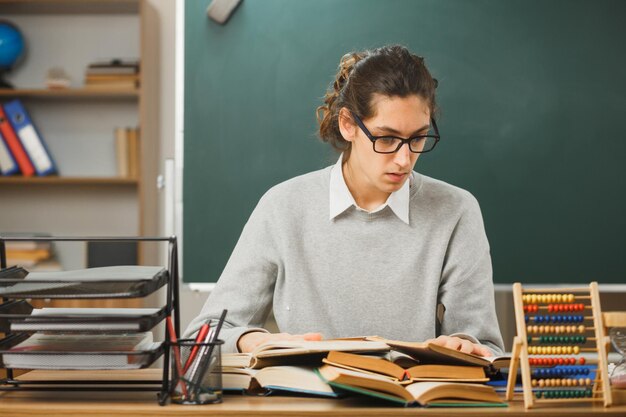 junger männlicher lehrer, der am schreibtisch mit schulwerkzeugen im klassenzimmer sitzt