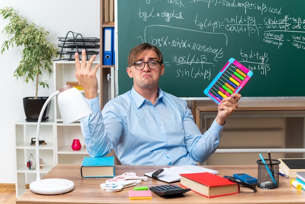 Junger männlicher Lehrer, der Brillen mit Rechnungen trägt, die verwirrt schauen, bereiten Lektion vor, die am Schulschreibtisch mit Büchern und Notizen vor Tafel im Klassenzimmer sitzt
