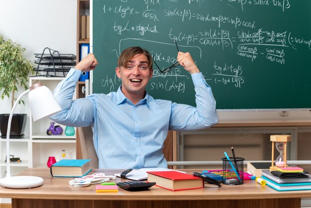 Junger männlicher Lehrer, der eine Brille hält, die glücklich und aufgeregt die Fäuste hebt, die an der Schulbank mit Büchern und Notizen vor der Tafel im Klassenzimmer sitzen