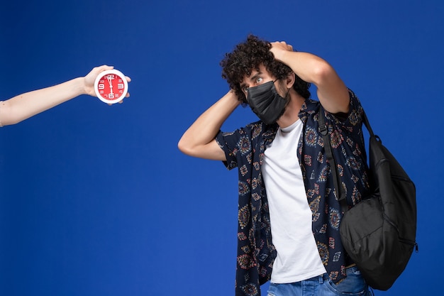 Junger männlicher Student der Vorderansicht, der schwarze Maske mit Rucksack auf hellblauem Hintergrund aufwirft.