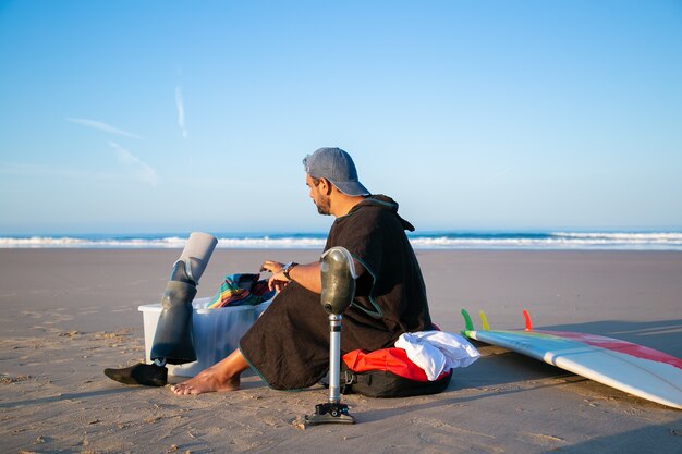 Junger männlicher Surfer, der nahe Brett am Strand sitzt und künstliche Gliedmaßen ändert