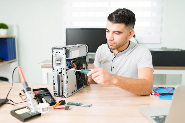 Junger männlicher Techniker, der für einen Kunden neue Komponenten auf einer defekten CPU installiert. Lateinischer Mann, der eine Reparaturwerkstatt betreibt