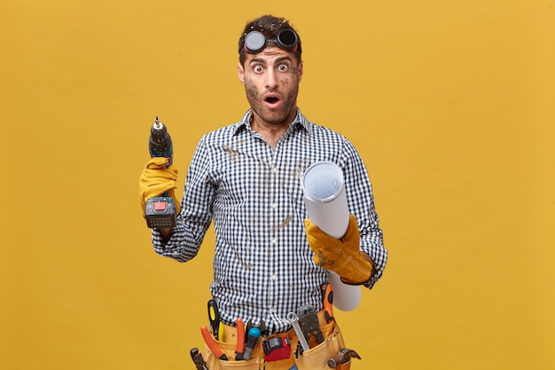 Kostenloses Foto junger männlicher techniker mit schmutzigem gesicht, das schutzhandschuhe trägt, die bohrmaschine und blaupause in seinen händen halten, die schockierten ausdruck erkennen, wie viel er tun sollte