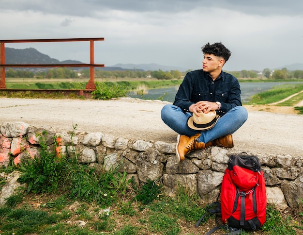 Junger männlicher Tourist, der mit Rucksack nahe schönem Fluss sitzt