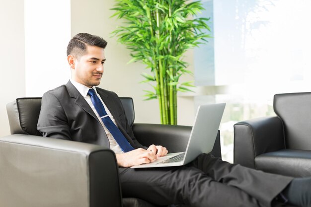 Junger männlicher Wirtschaftsanalytiker, der auf einem Laptop-Computer im Internet surft, während er auf einem Stuhl in der Lobby des Büros sitzt