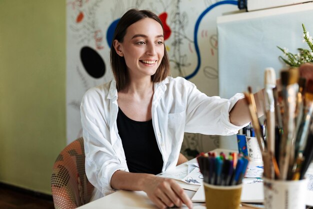 Junger Maler am Schreibtisch wählt freudig Pinsel mit großer Musterleinwand auf Hintergrund zu Hause aus