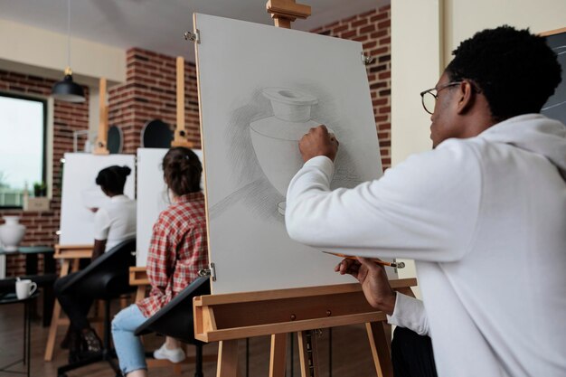 Junger Maler sitzt vor weißer Leinwand und zeichnet Vasenmodell mit grafischem Bleistift, der während des Kunstunterrichts an Skizzenillustration arbeitet. Multiethnisches Team nimmt an Skizzierunterricht im Kreativitätsstudio teil