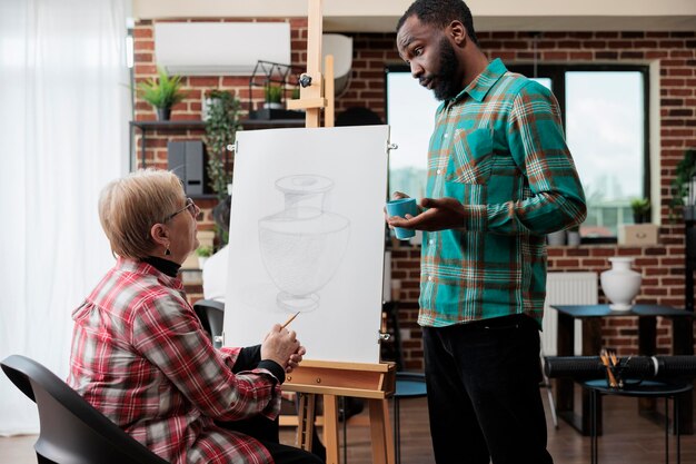 Junger Malerlehrer erklärt älteren Frauen, die bei der grafischen Auslosung im Kreativitätsstudio zusammenarbeiten, Skizzenillustration. Diverse Teams zeichnen Vasenmodelle auf Leinwand und lernen neue künstlerische Fähigkeiten