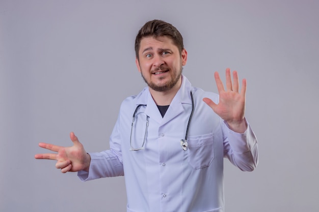 Junger Mann Arzt, der weißen Kittel und Stethoskop trägt, das frustriert aussieht und Nummer acht mit Fingern und Händen zeigt