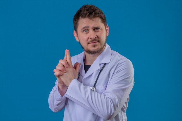 Junger Mann Arzt, der weißen Mantel und Stethoskop hält symbolische Waffe mit Handgeste über lokalisiertem blauem Hintergrund hält