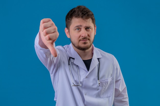 Junger Mann Arzt trägt weißen Kittel und Stethoskop zeigt Daumen nach unten, zeigt Abneigung über isolierten blauen Hintergrund