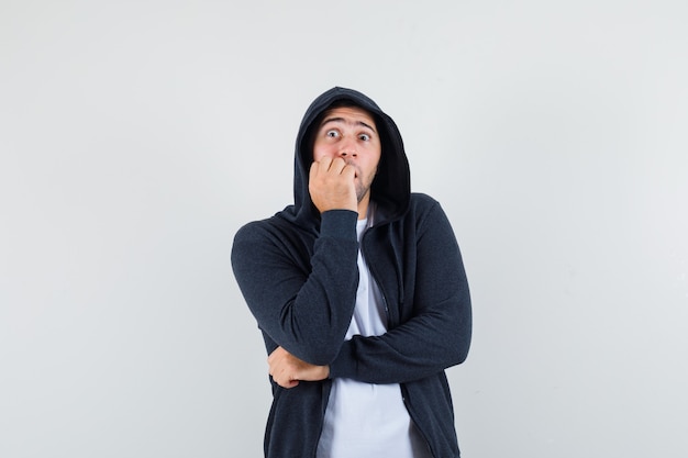 Junger Mann beißt Faust emotional in T-Shirt, Jacke und ängstlich aussehend. Vorderansicht.