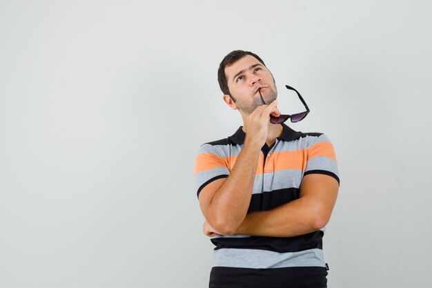 Junger Mann beißt seine Brille, während er in T-Shirt, Sonnenbrille und nachdenklich schaut. Platz für Text