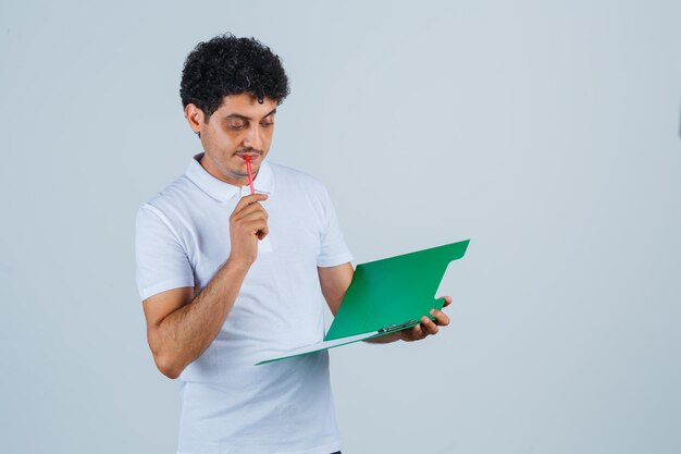 Junger Mann beißt Stift, hält Notizbuch und sieht es in weißem T-Shirt und Jeans an und sieht nachdenklich aus. Vorderansicht.