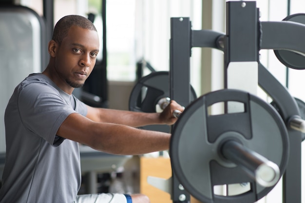 Junger Mann, der am Barbell steht und Kamera betrachtet