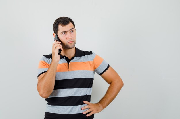 Junger Mann, der am Telefon spricht, während er im T-Shirt beiseite schaut und konzentriert schaut. Vorderansicht.