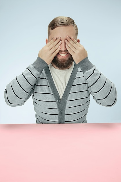 Kostenloses Foto junger mann, der am tisch im studio mit geschlossenen augen lokalisiert auf blau sitzt. ich kann nichts konzept sehen. minimalismus-stil