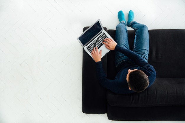 Junger Mann, der an dem Laptop, sitzend auf schwarzem Sofa, weißer Boden arbeitet.