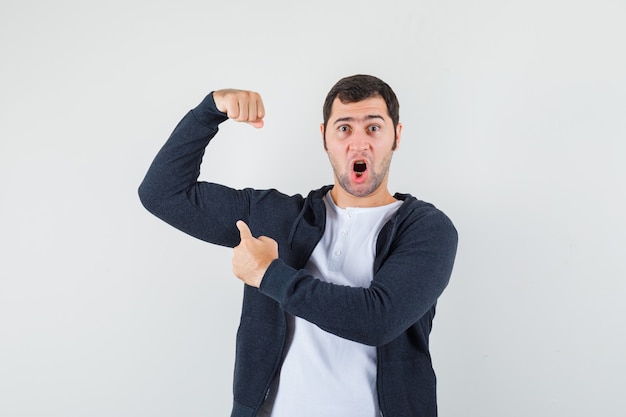 Junger Mann, der auf Armmuskeln im T-Shirt, in der Jacke und in der kraftvollen Vorderansicht zeigt.