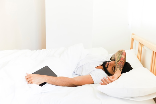 Junger Mann, der auf Bett mit digitaler Tablette schläft